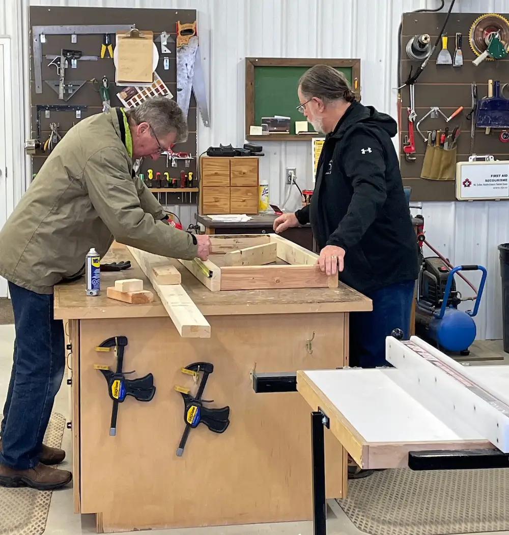 Two men working on a woodworking project. Tools in the background.